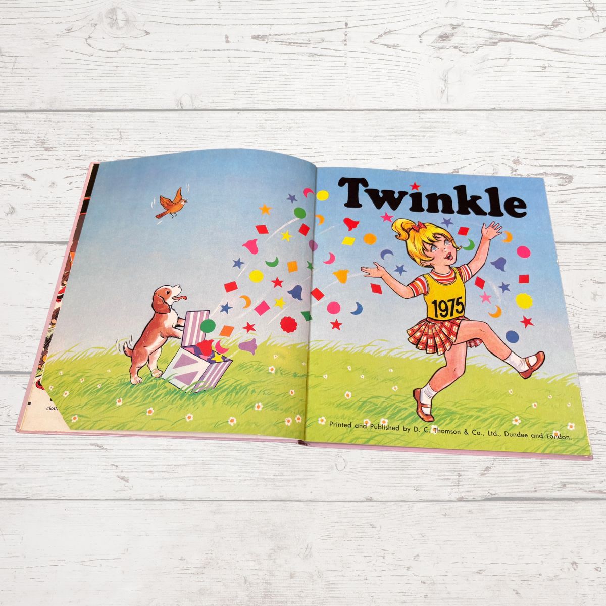 An open vintage children's book titled 'Twinkle' laid flat on a white wood plank surface. The illustrated spread shows a girl running with colorful confetti and stars, a dog beside a gift box, and a grassy field under a blue sky.