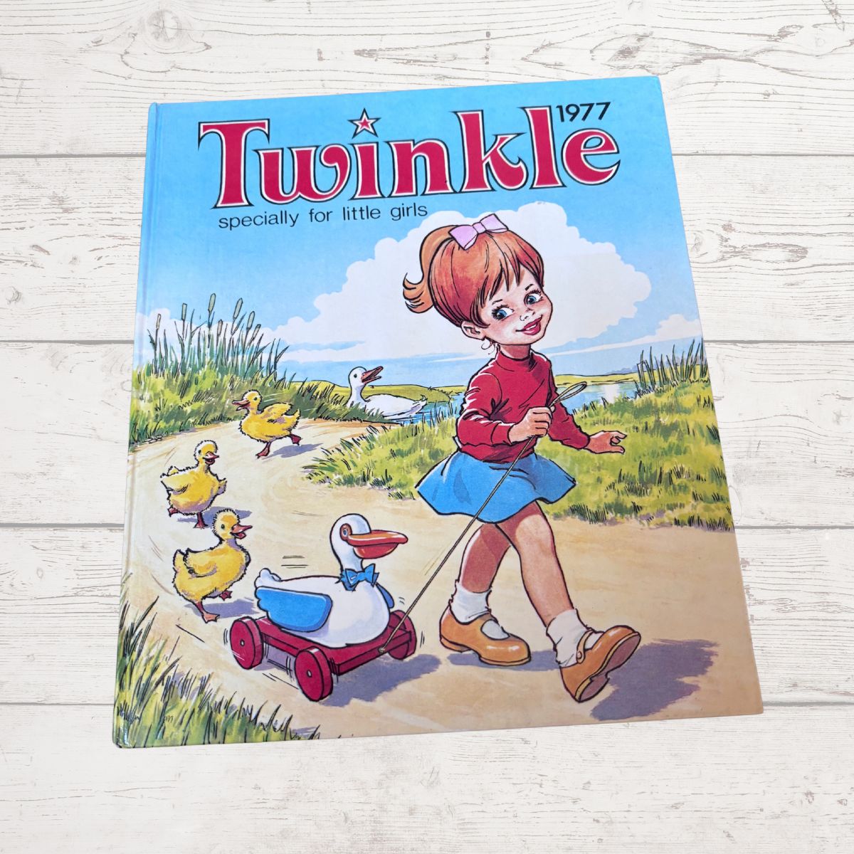 A vintage 1977 children's book titled 'Twinkle' lies flat on a white wooden surface. The cover features colorful cartoon artwork of a girl in a red top and blue skirt pulling a duck in a red wagon along a path, with other ducks in the background.