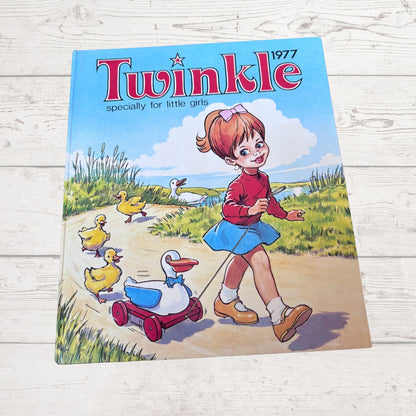 A vintage 1977 children's book titled 'Twinkle' lies flat on a white wooden surface. The cover features colorful cartoon artwork of a girl in a red top and blue skirt pulling a duck in a red wagon along a path, with other ducks in the background.