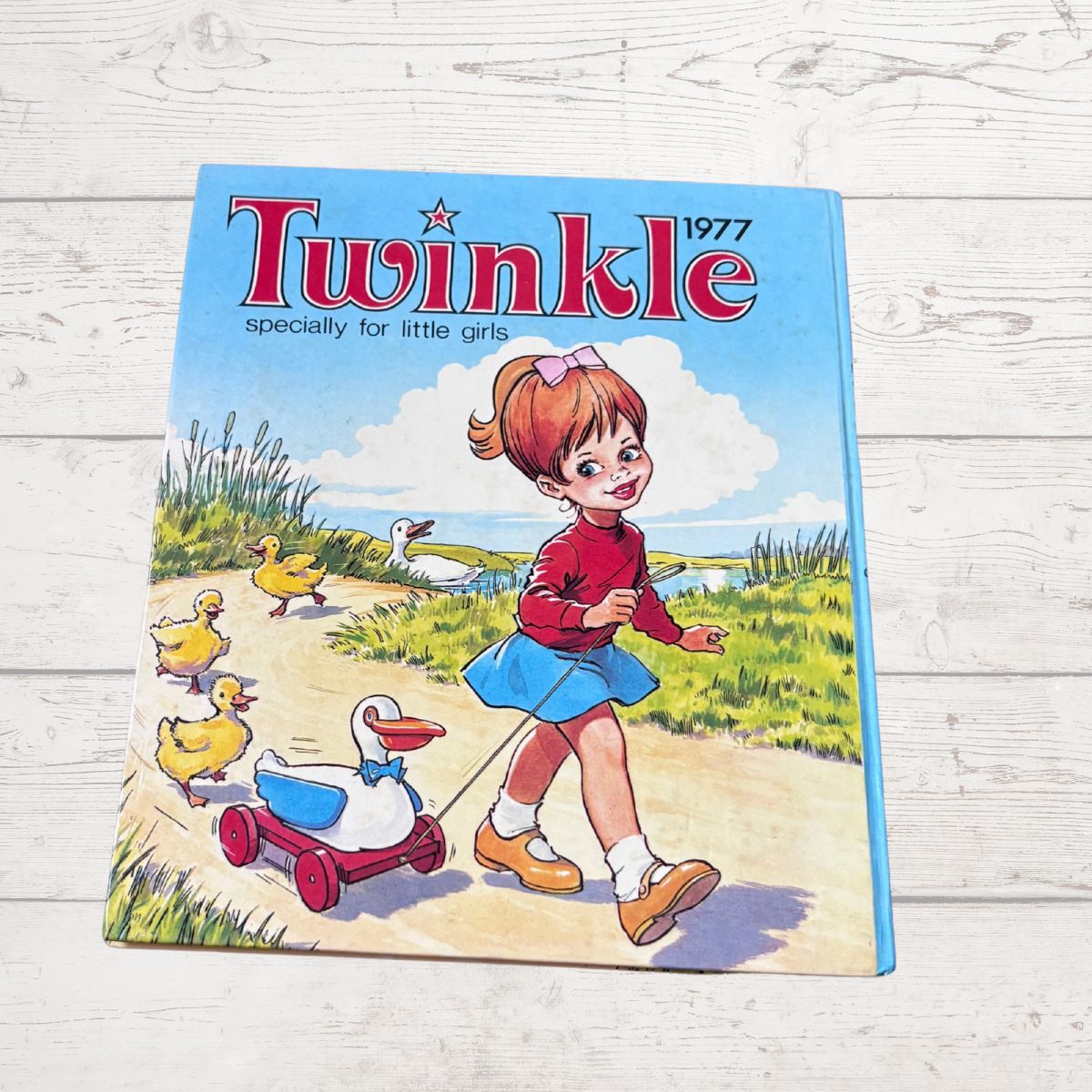 A vintage 1977 Twinkle magazine for girls, featuring a colorful illustration of a girl walking with ducks, displayed on a light-colored wooden background.