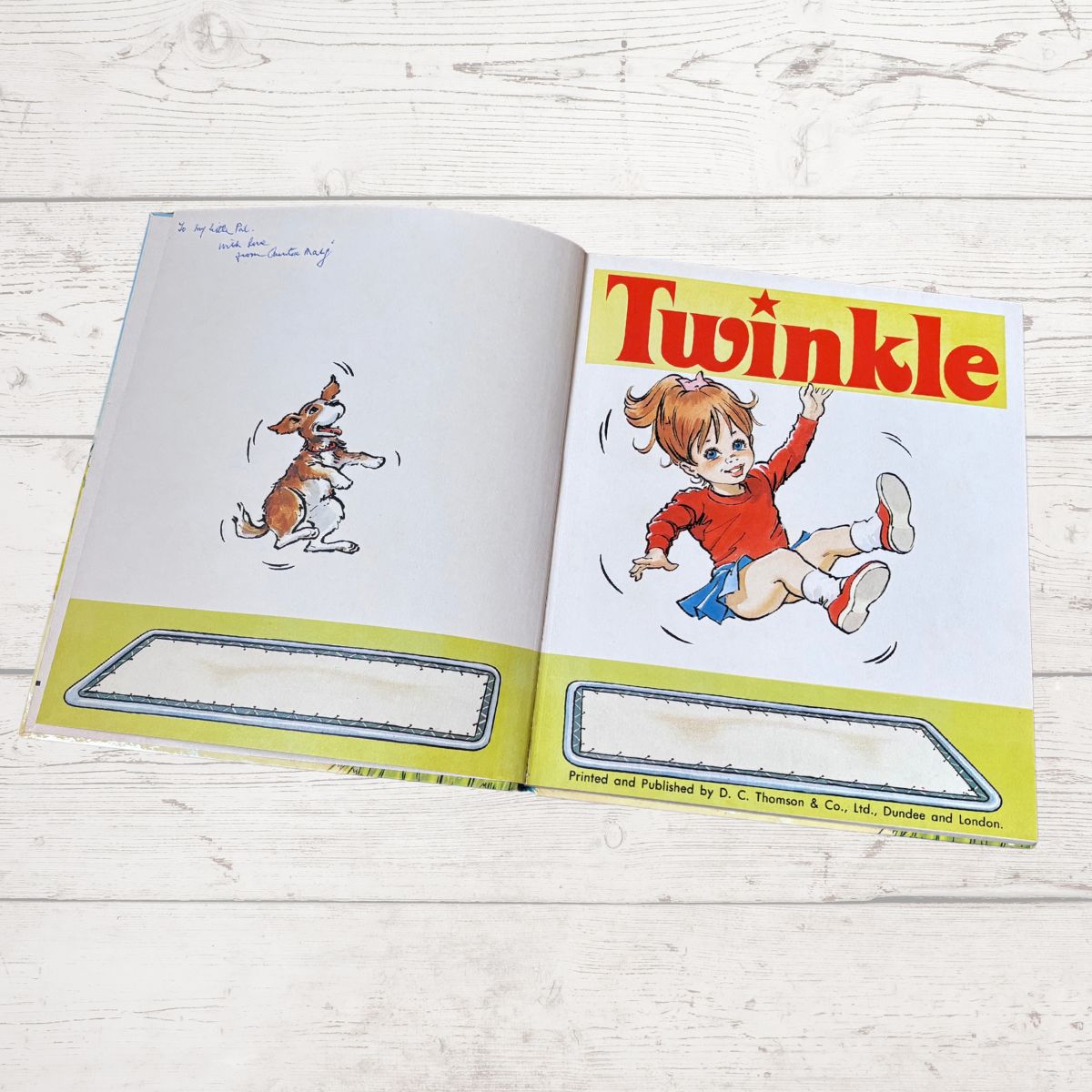 An open vintage children's book titled 'Twinkle' laid flat on a light whitewashed wooden surface. The right page shows a colorful cartoon illustration of a girl named Twinkle with the title in large red letters, and the left page shows a small sketch of a dog.
