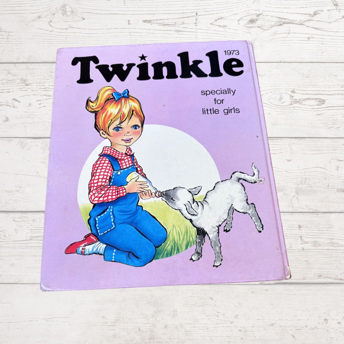 A vintage 1973 children's book titled 'Twinkle' for little girls, featuring a purple cover with an illustration of a girl in blue overalls kneeling beside a white lamb. The book is photographed on a light-colored wooden surface.