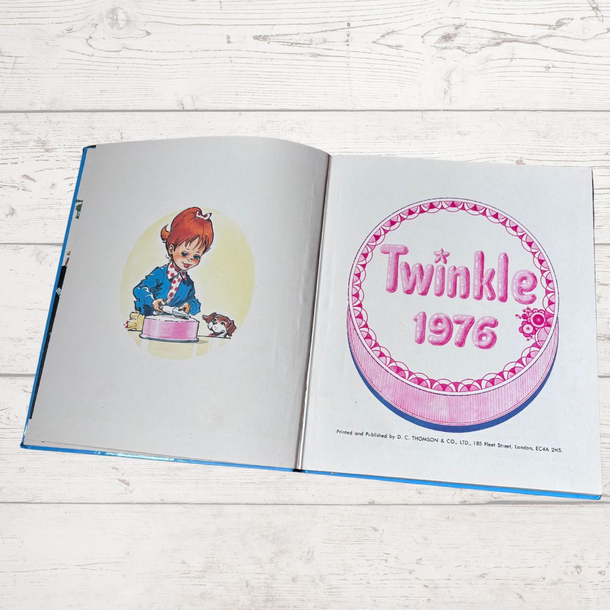 An open vintage book lying on a white wood surface, showing illustrated pages. The right page features a large pink-and-white Twinkle 1976 cake illustration, and the left page shows a cartoon character drawing.