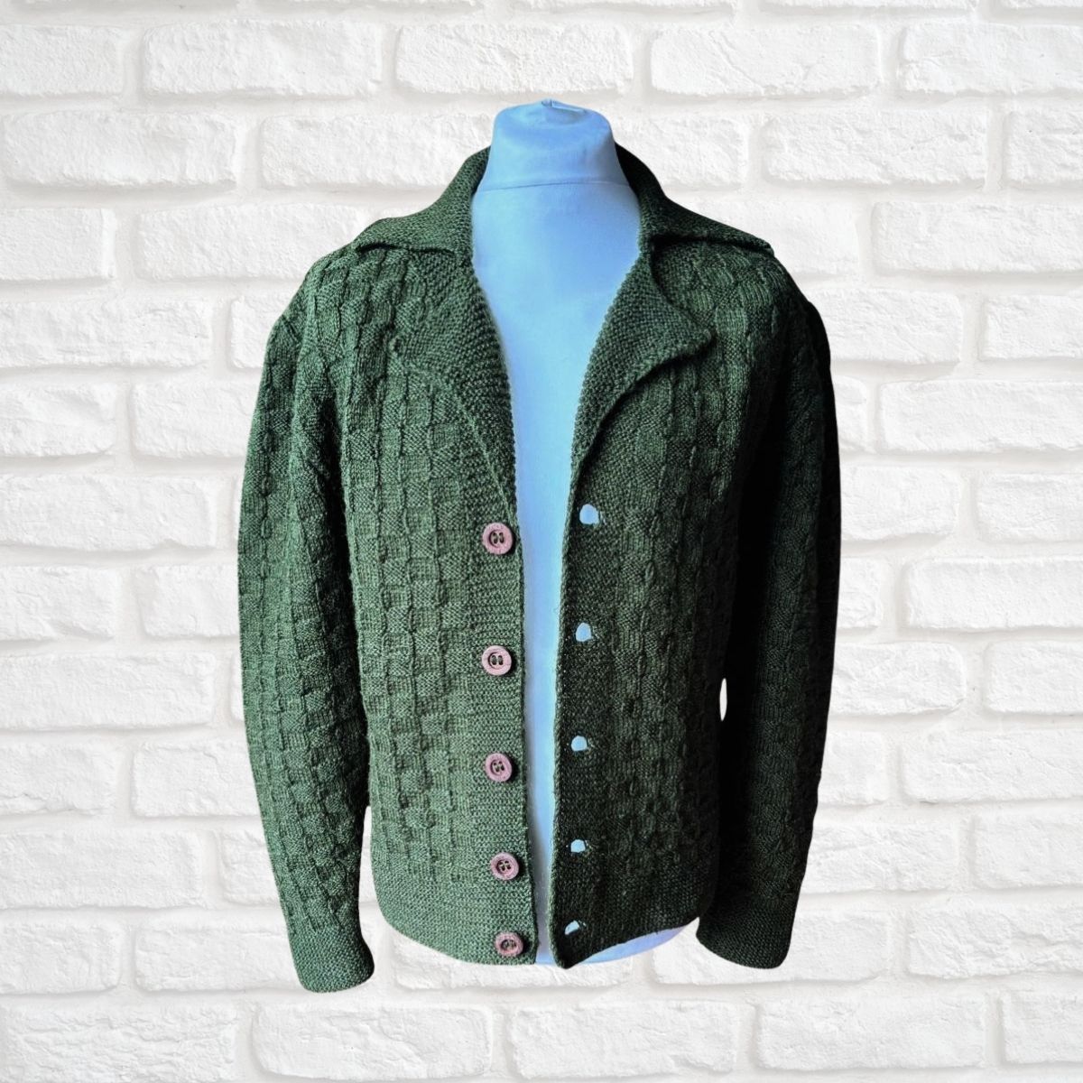 A dark green cable-knit cardigan sweater displayed on a light blue mannequin torso against a white painted brick wall. The cardigan features a textured knit pattern, a notched collar, and pink buttons.