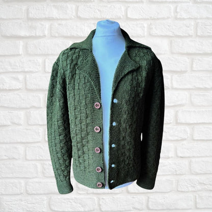 A dark green cable-knit cardigan sweater displayed on a light blue mannequin torso against a white painted brick wall. The cardigan features a textured knit pattern, a notched collar, and pink buttons.