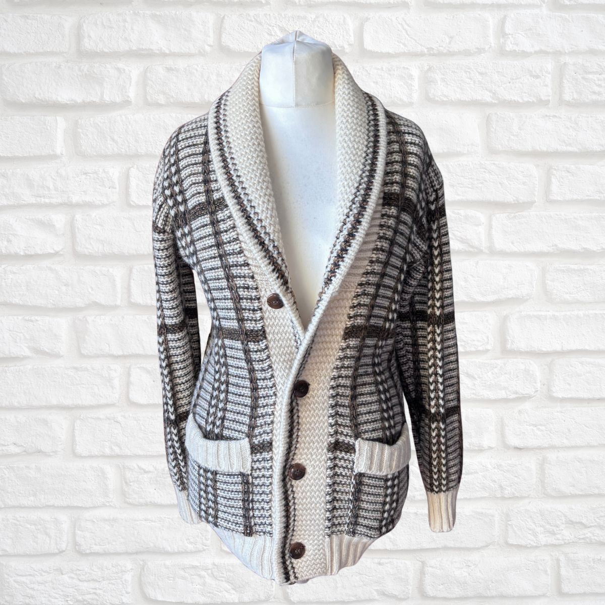A unisex knit cardigan sweater with a shawl collar, displayed on a white mannequin torso against a white brick wall. The cardigan features a plaid and geometric pattern in black, gray, and cream, with dark brown buttons.