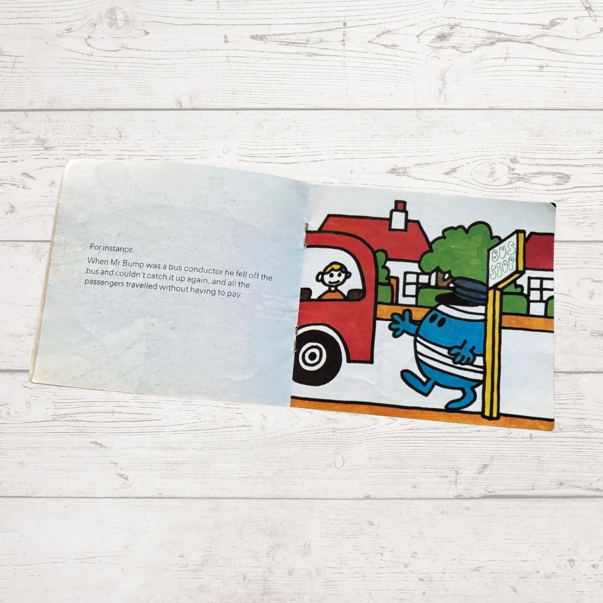 Open book with a colorful illustration of a character Mr Bump near a bus stop on a wooden surface