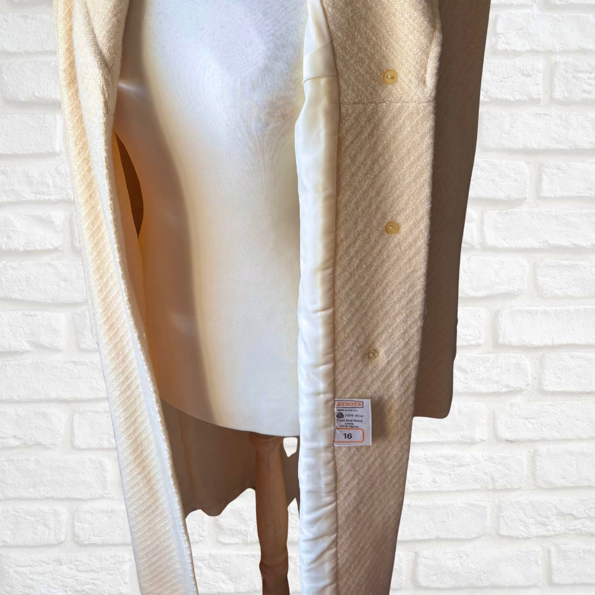Beige wool coat on a mannequin against a white brick wall.
