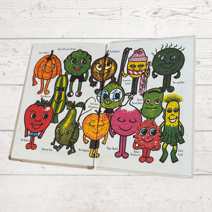 Open book with colorful illustrations of fruit and vegetable characters on a wooden surface.