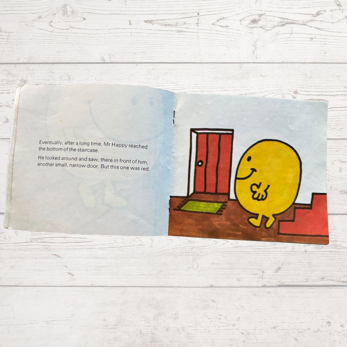 Mr Happy next to a red door at the bottom of a staircase 