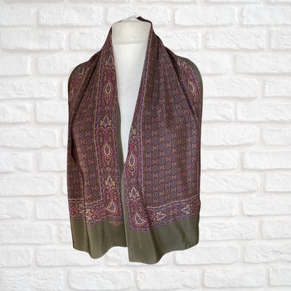 A long length vintage green silk paisley print scarf displayed draped on a mannequin against a brick wall backdrop.