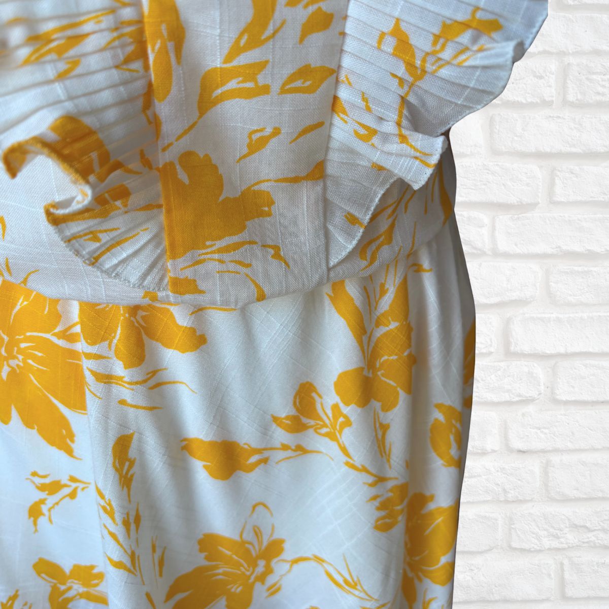 Yellow floral patterned dress  with ruffled edge against a white brick wall.