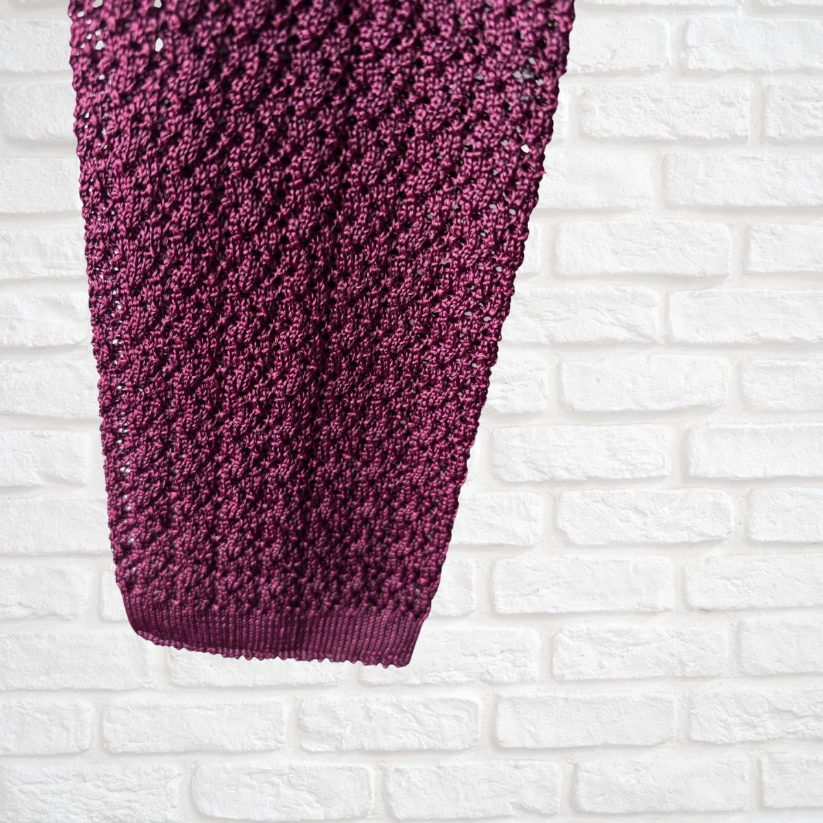 Close-up of a textured burgundy neck tie against a white brick wall.