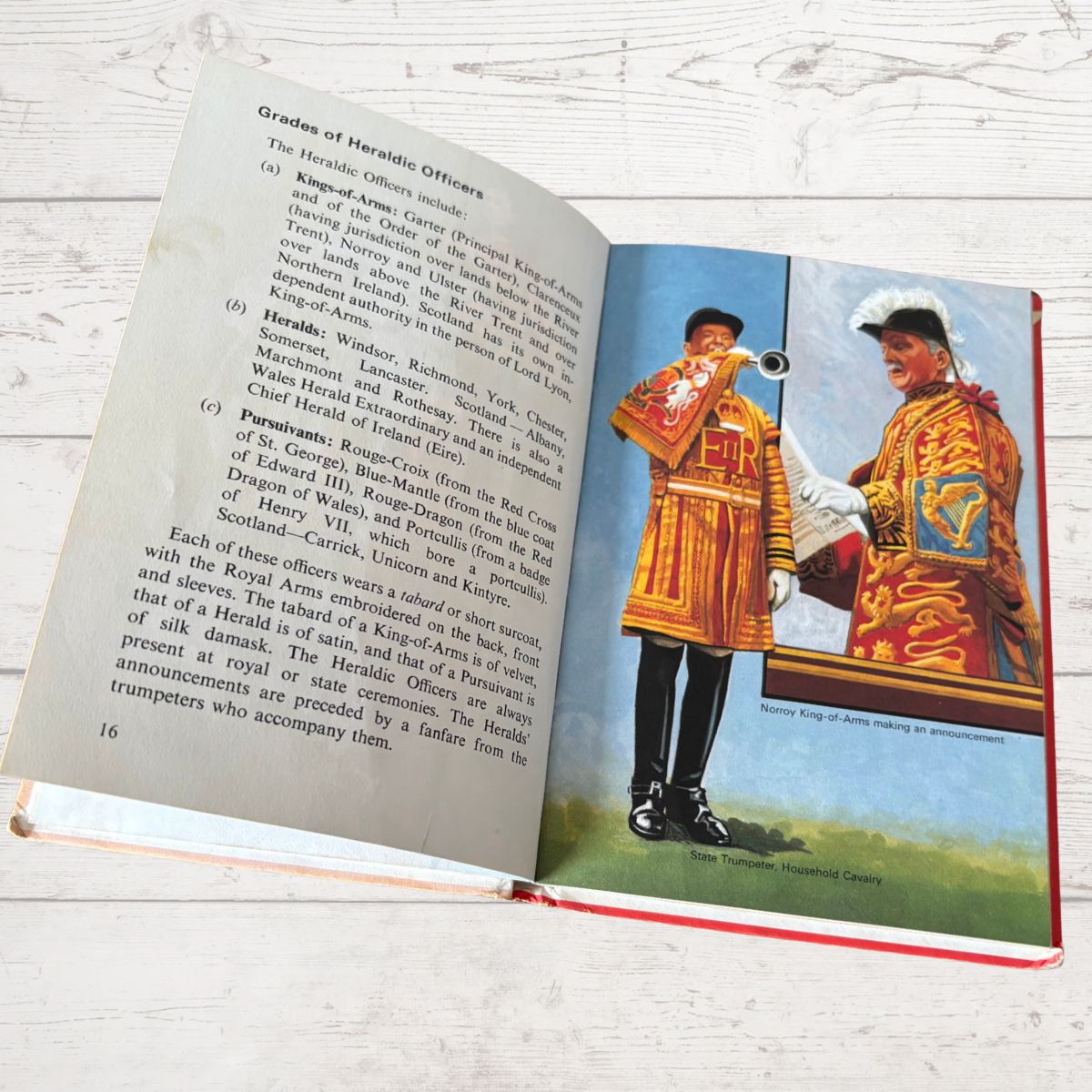 Open book with illustrations of heraldic officers and text on a wooden surface