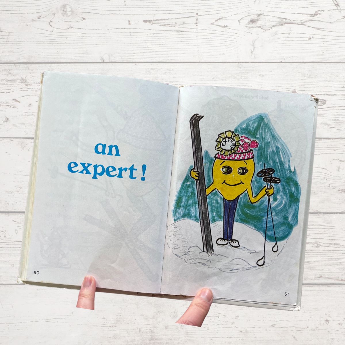 Open book with a cartoon character holding skis and snowshoes, labeled 'an expert!' on a wooden surface.