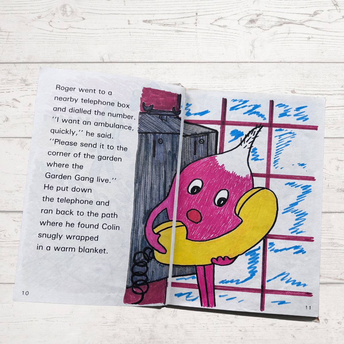 Open book with a colorful illustration of a pink character with a yellow beak, standing next to a telephone booth on a wooden surface.