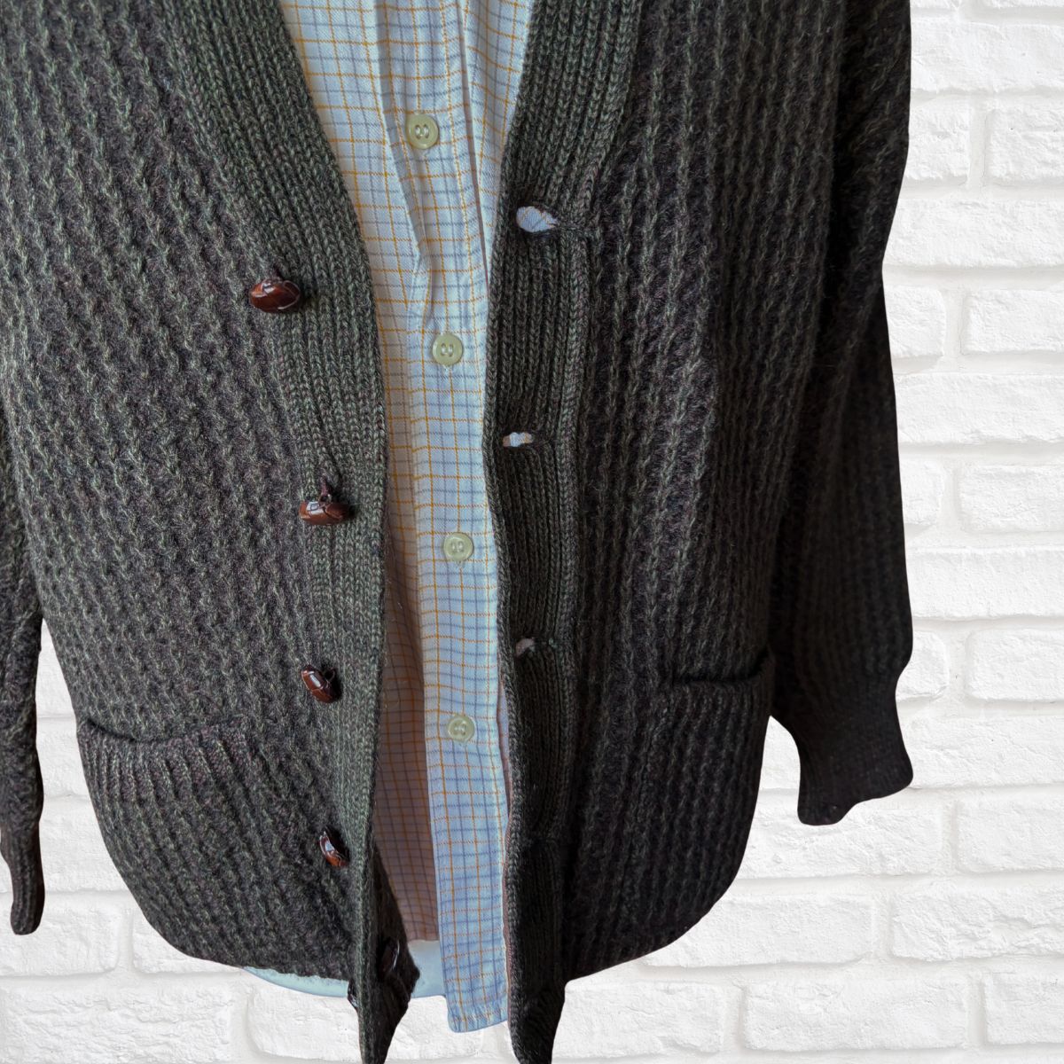 A wool cardigan over a plaid shirt against a white brick wall.