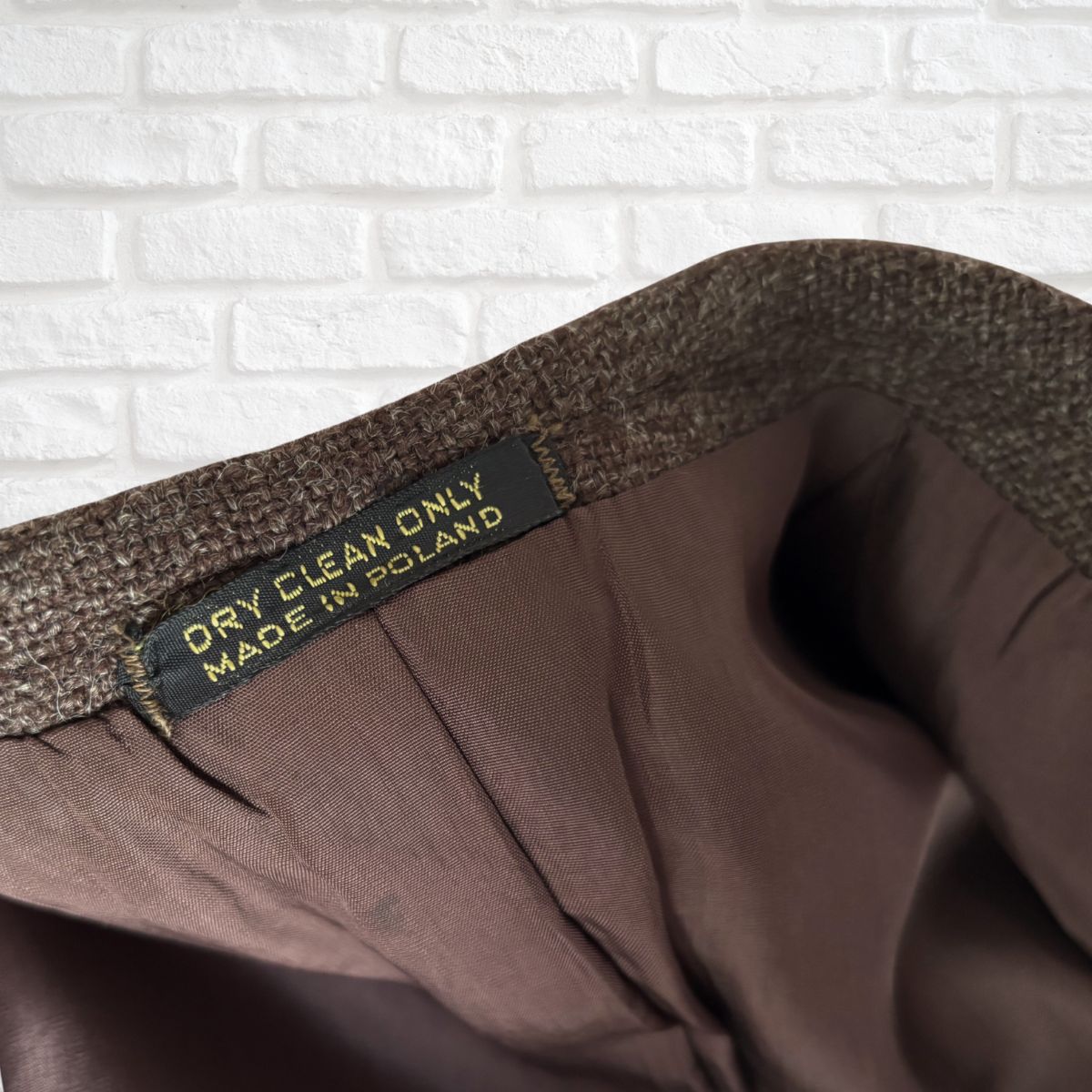 Brown fabric with a label indicating 'Dry Clean Only, Made in Poland' against a white brick wall background.