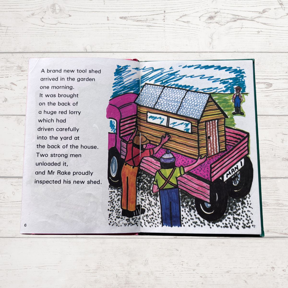 Illustrated book page with a cartoon of a truck carrying a shed and people around it, on a wooden surface.