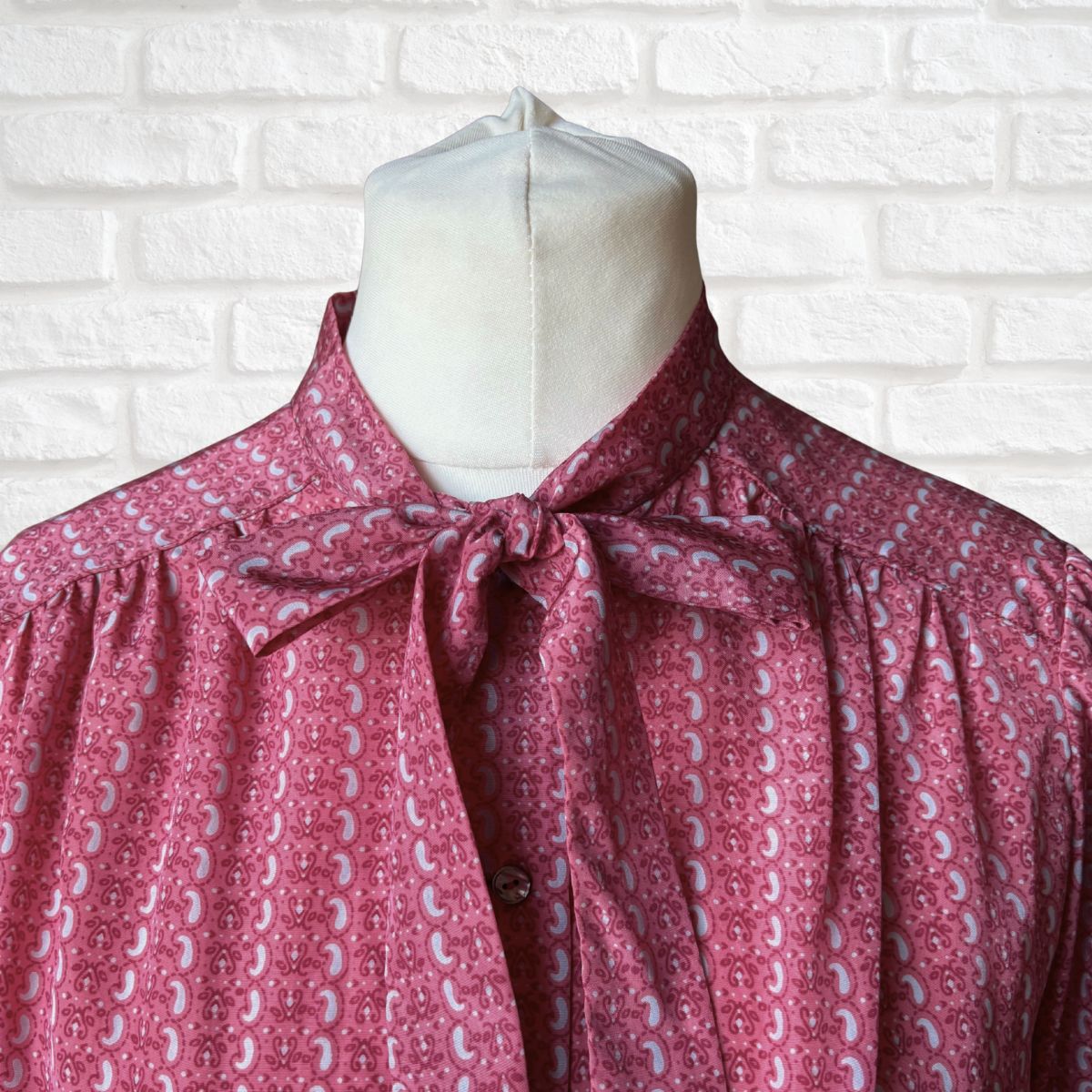 Mannequin wearing a pink patterned blouse against a white brick wall.