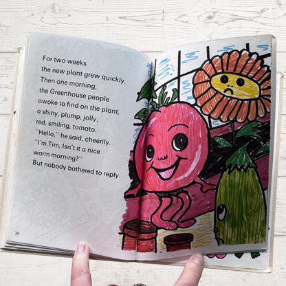 Open book with a colorful illustration of a tomato character and a sunflower on a white wooden surface.