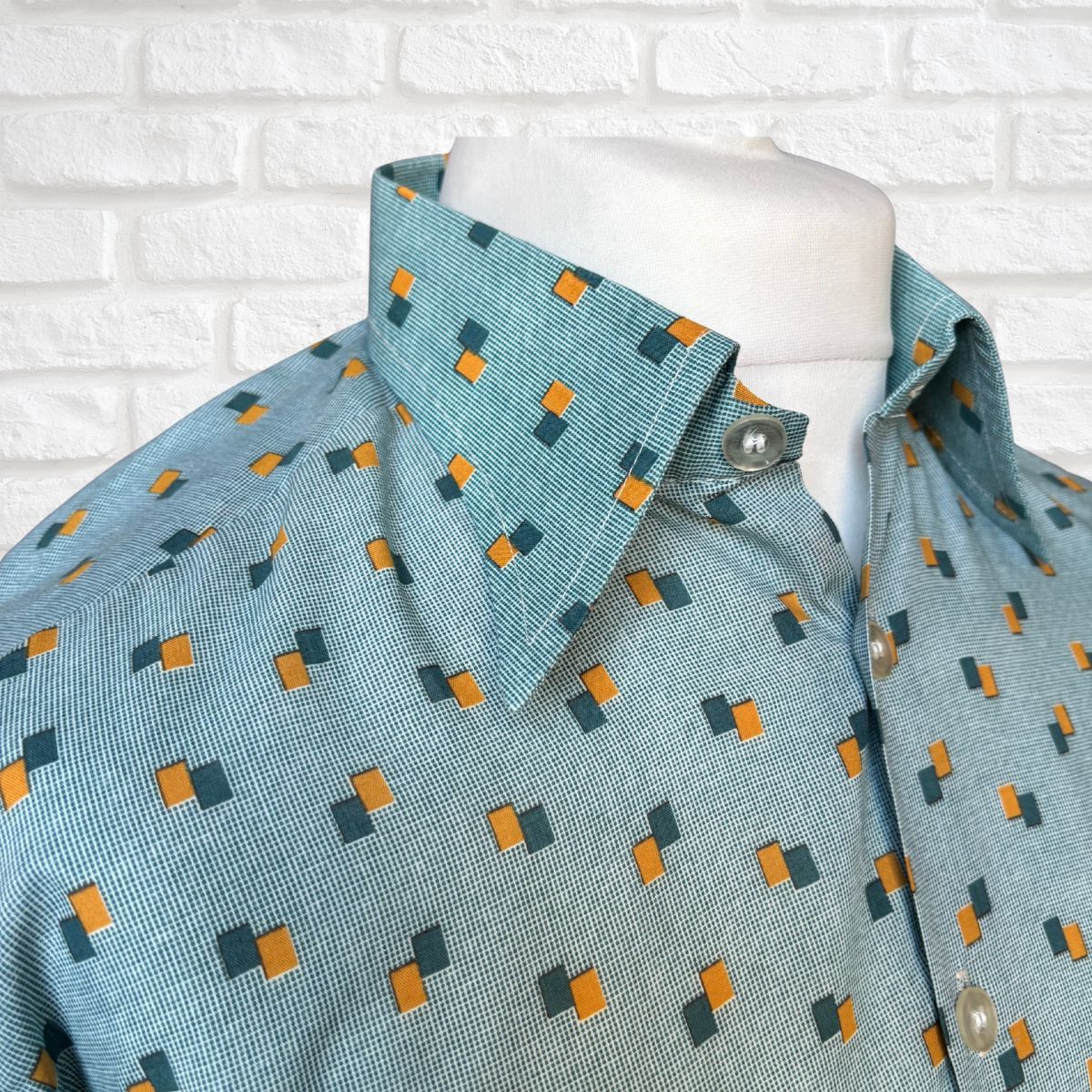 Mannequin wearing a teal shirt with geometric pattern against a white brick wall.