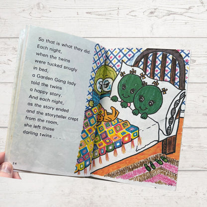 Open book with illustrated page of two green characters in bed, colorful blanket, and text on a wooden surface.