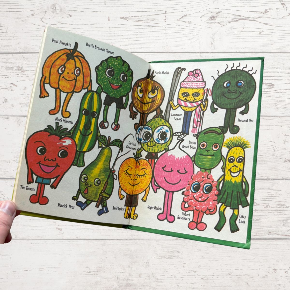 Illustrated book open to a page with colorful vegetables and fruits with faces on a white wooden surface.