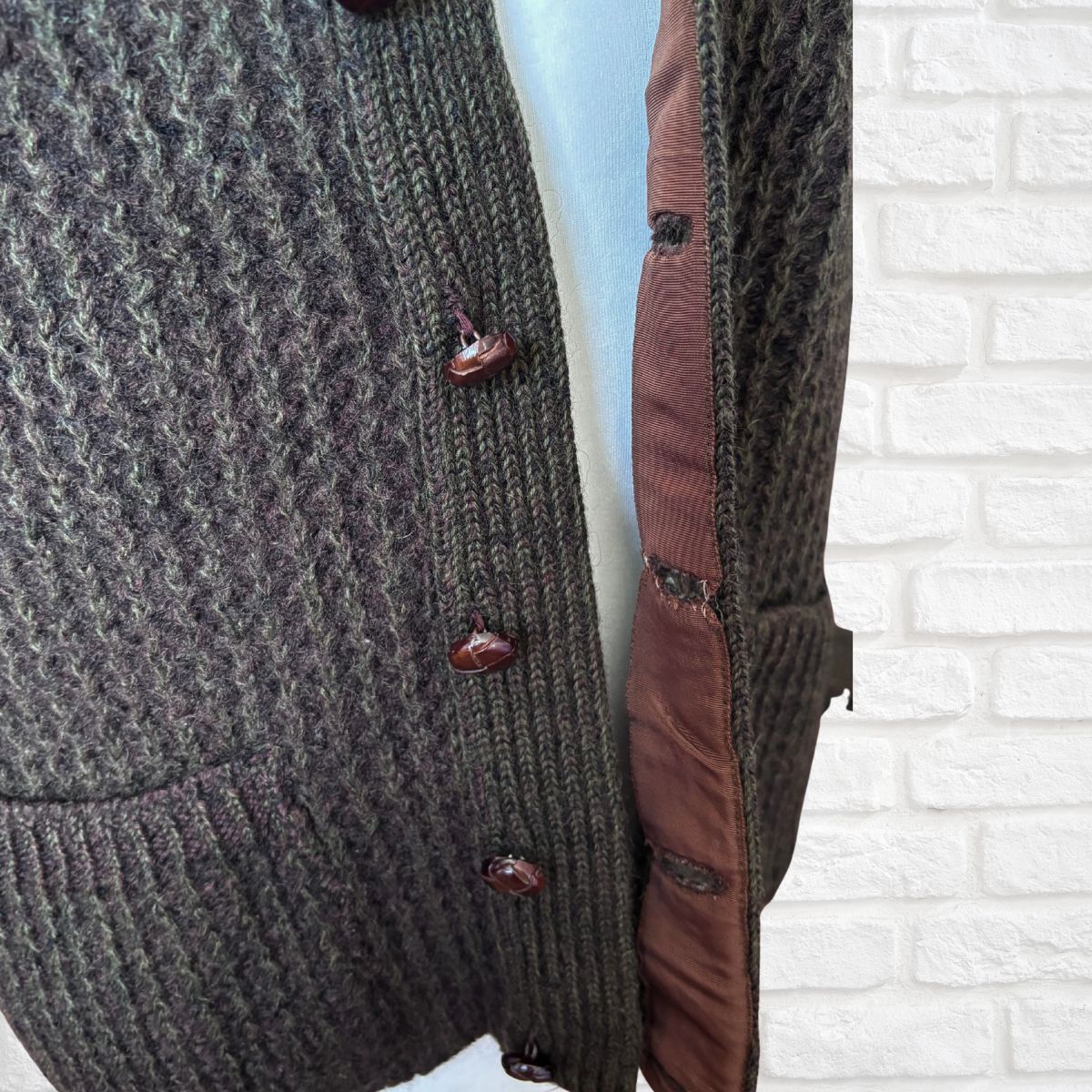 Close-up of a textured cardigan with brown buttons against a white brick wall.