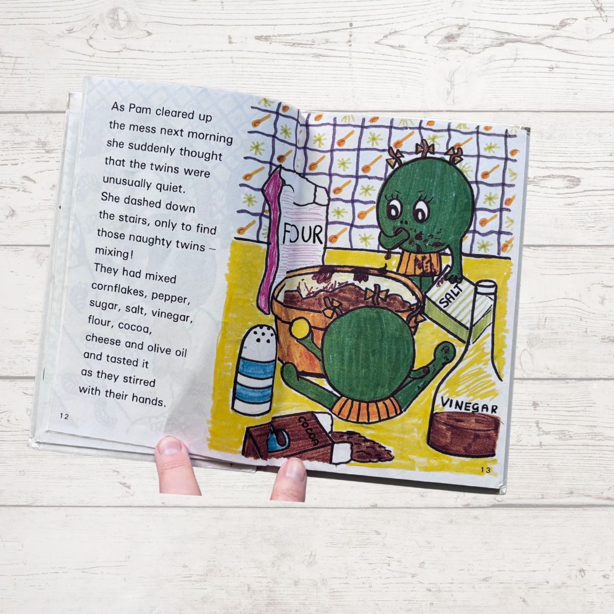 Open book with a colorful illustration of a green creature in a kitchen, held by a hand on a light wooden surface.