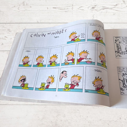 Open book showing a comic strip of Calvin and Hobbes on a light wooden surface.