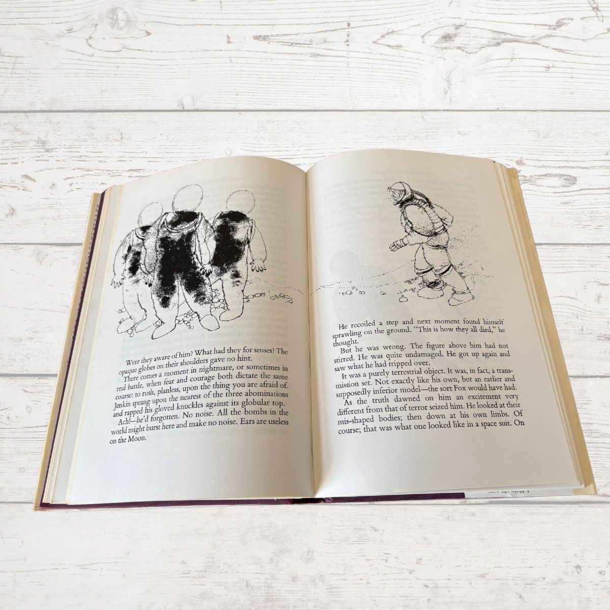 An open vintage hardcover book lying flat on a white wooden surface, showing illustrated pages with black ink drawings and printed text.