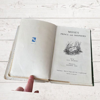 Open book titled 'Moses Prince and Shepherd' held by a hand on a light wooden surface.