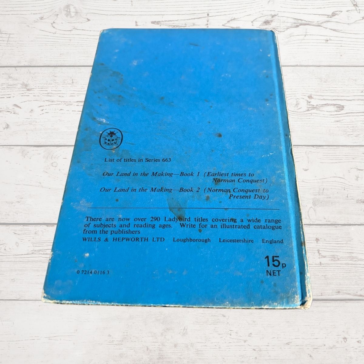 A vintage blue book cover photographed from above on a white painted wood surface. The back cover shows printed text, a small publisher logo, and the price '15p NET'.