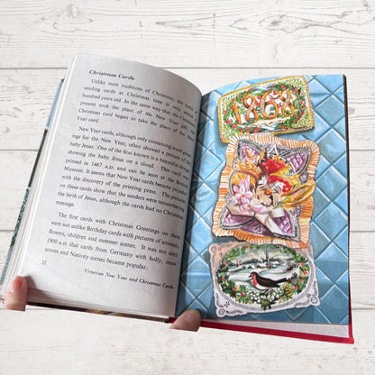 An open book showing printed text on the left page and a colorful vintage-style Christmas card illustration on the right page, held by two hands over a white wood surface.