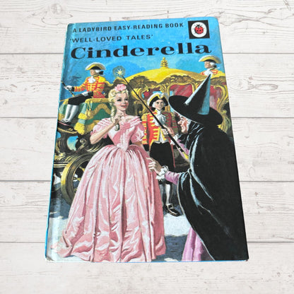 Vintage paperback book titled 'Cinderella' from the 'A Ladybird Easy-Reading Book' series, featuring a colorful cover illustration of Cinderella in a pink gown and the witch in black, placed on a light wood surface.
