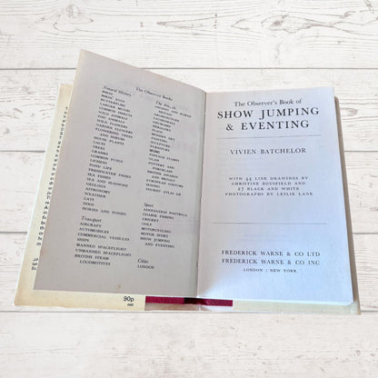 An open vintage book titled 'The Observer's Book of Show Jumping & Eventing' by Vivien Batchelor, displayed on a light whitewashed wooden surface. The left page shows a list of jumpers, and the right page displays the title page with publisher details for Frederick Warne & Co Ltd.