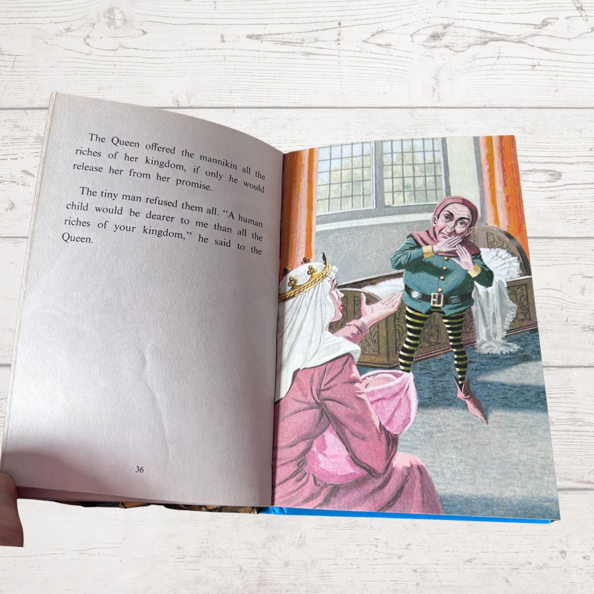 An open children's book lying on a light wood surface, showing a printed illustration of a queen in pink clothing talking to a small fairy in a green outfit. A hand is visible holding the left edge of the book.
