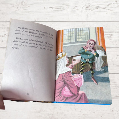 An open children's book lying on a light wood surface, showing a printed illustration of a queen in pink clothing talking to a small fairy in a green outfit. A hand is visible holding the left edge of the book.