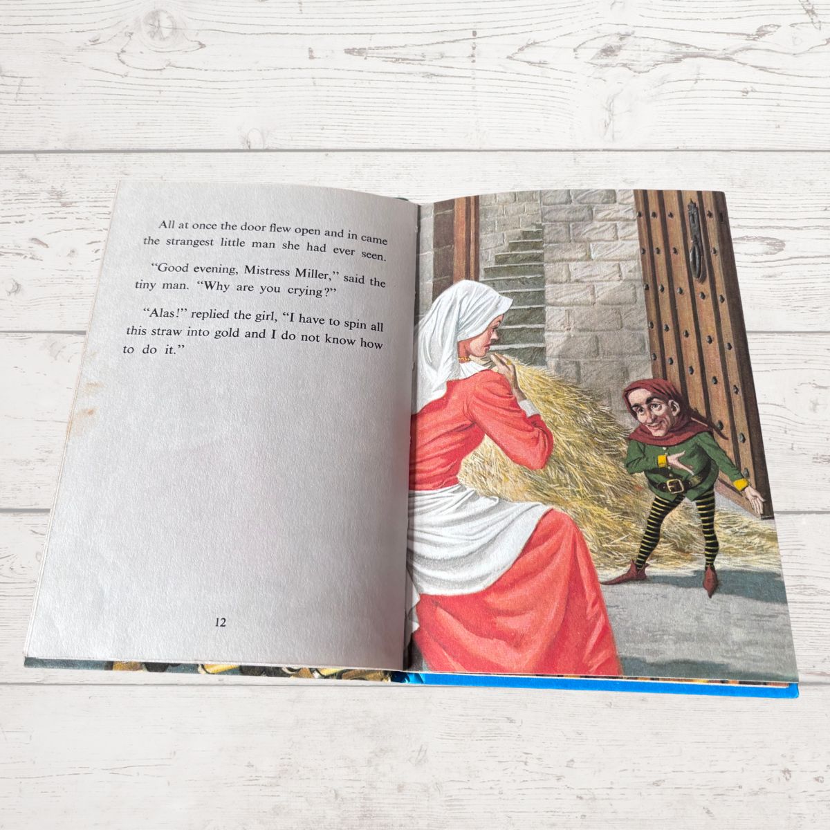 An open children's book lying on a white painted wood surface, showing a printed story page with text on the left and an illustrated scene on the right featuring a woman in a red dress and a small elf-like figure.