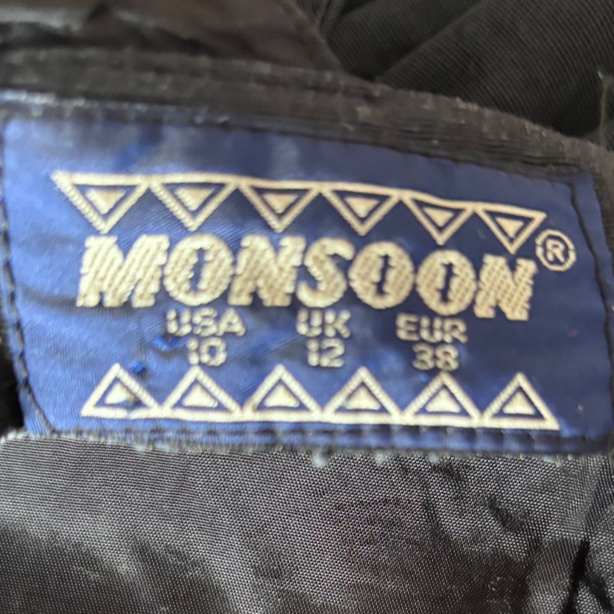 Close-up of a Monsoon garment label sewn onto black fabric, showing the brand name in gold embroidery on a blue background with size conversions.