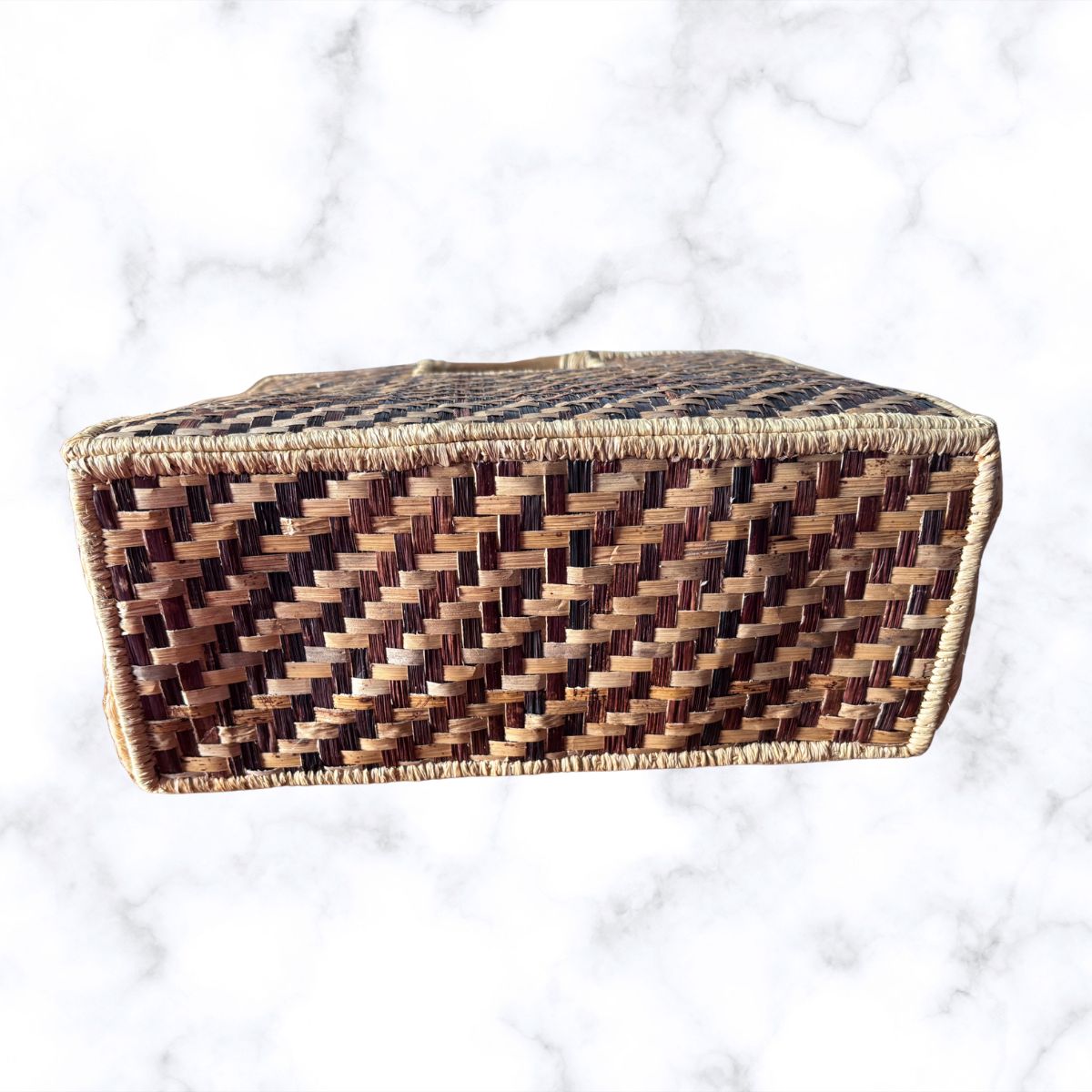 A rectangular woven basket with a diamond-patterned weave in shades of brown and beige, set against a white marble background.