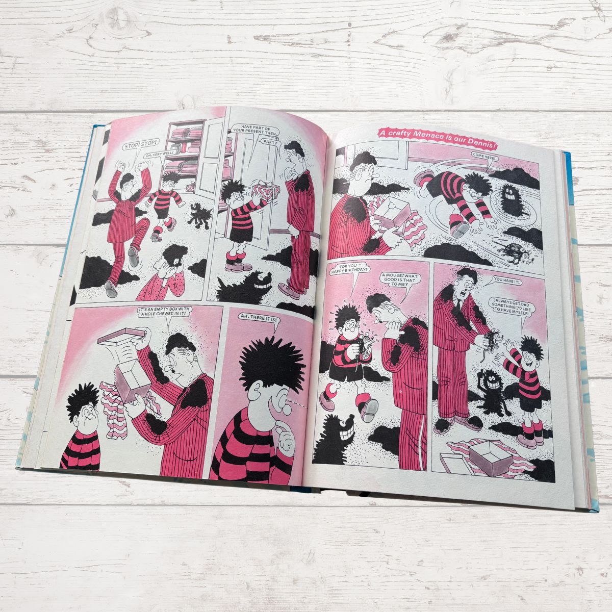 An open illustrated comic book laid on a white wood surface, showing pink, black, and white cartoon panels with multiple characters and speech bubbles.
