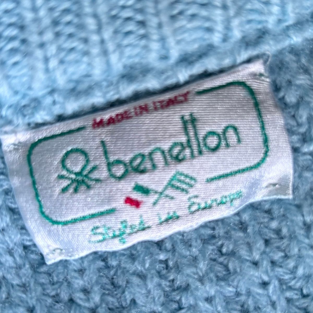 A close-up of a light blue knit fabric tag with a white sewn-in label reading 'benetton' in green, 'MADE IN ITALY' in red, and 'Stafet in Europa' in green, with small red and green flag details.