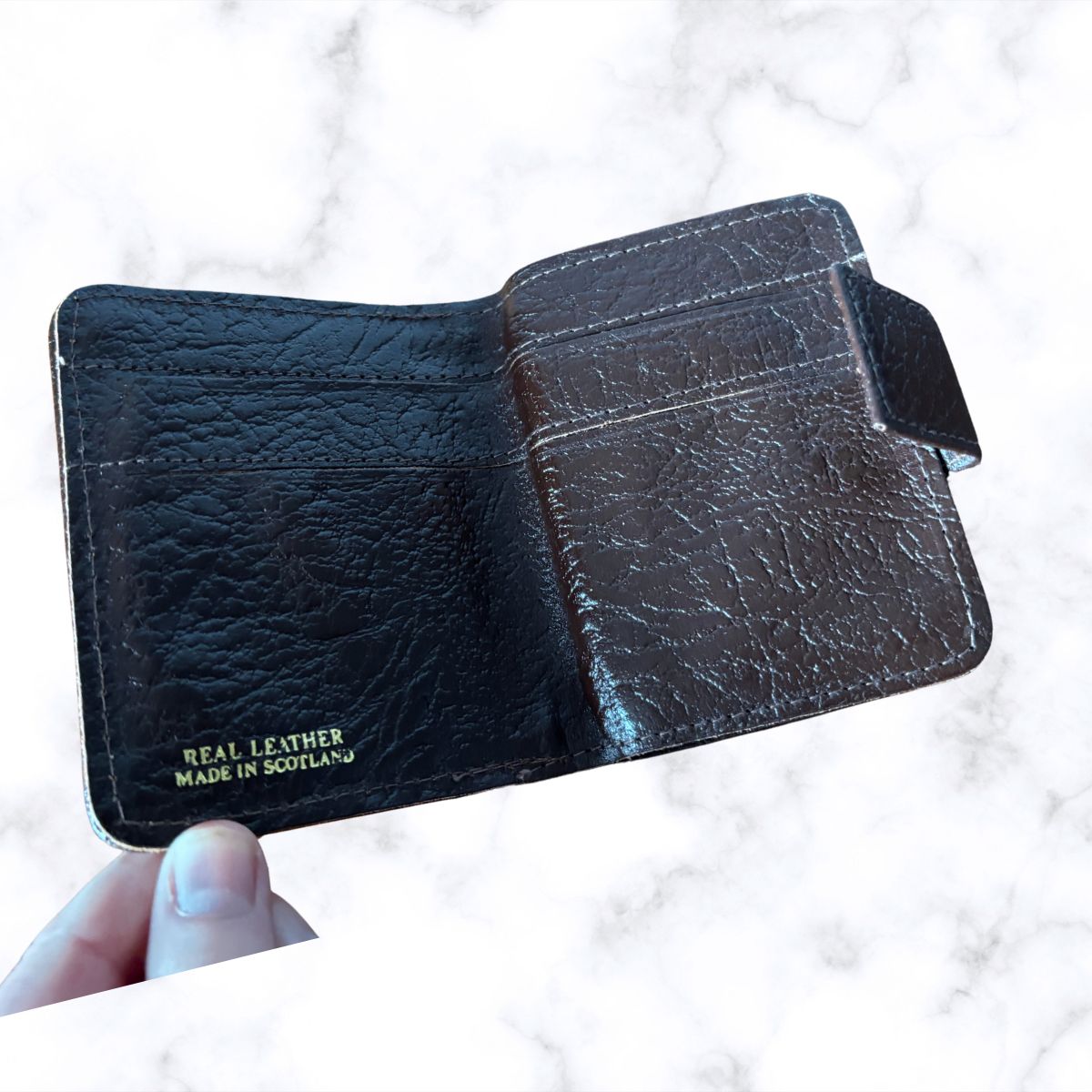 Black leather wallet with 'Real Leather Made in Scotland' text held by a hand on a light background