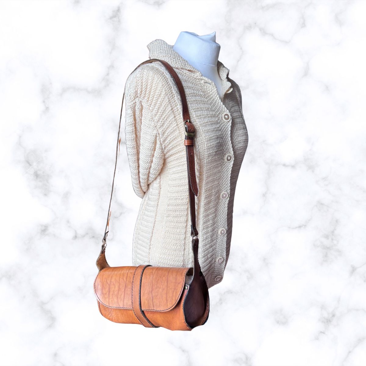 A brown leather shoulder bag with a textured finish and dark brown trim, displayed on a cream-colored knit cardigan sweater on a mannequin torso against a white marble background.