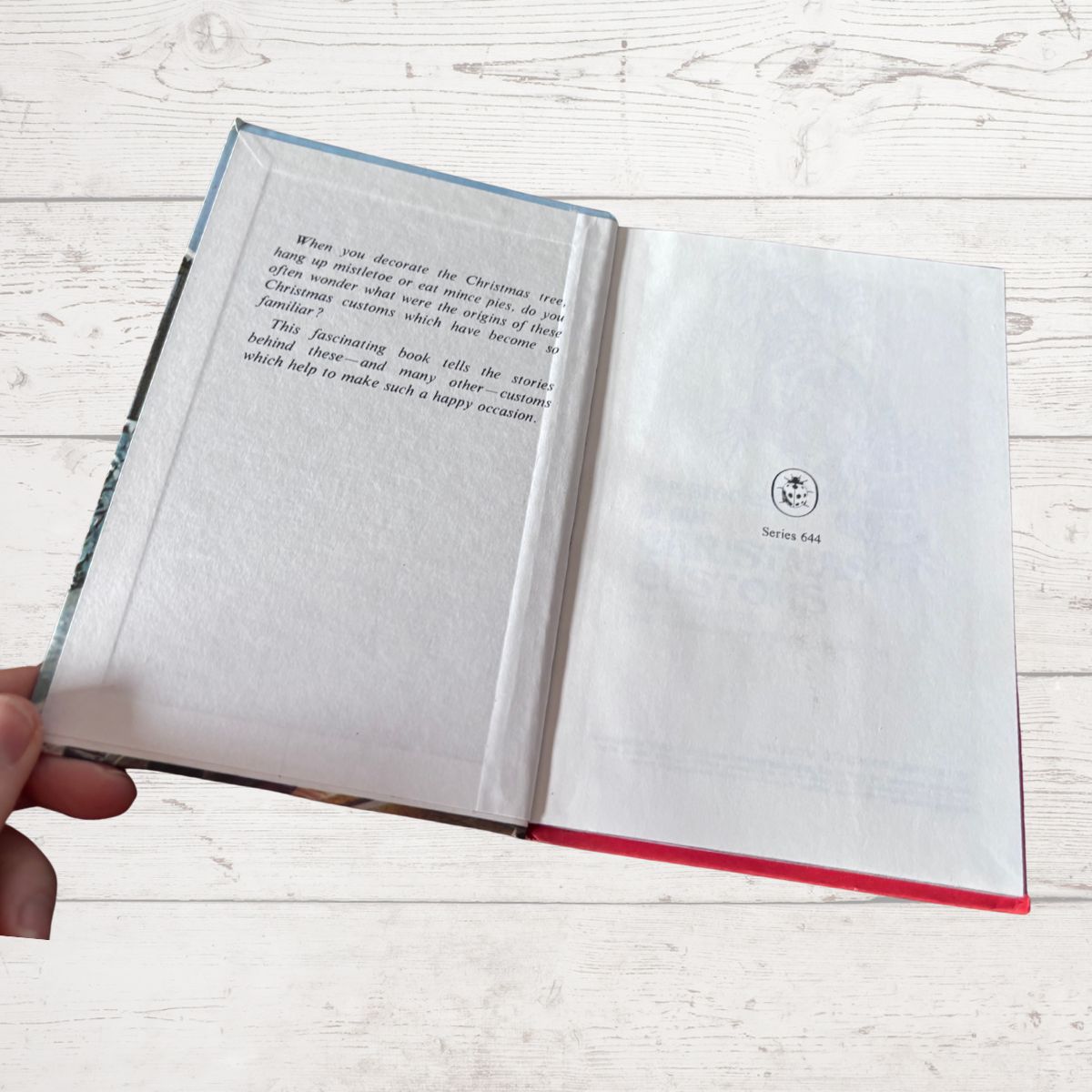 An open hardcover book with printed text on the left page and a blank page on the right, held by a hand against a light wood plank background.