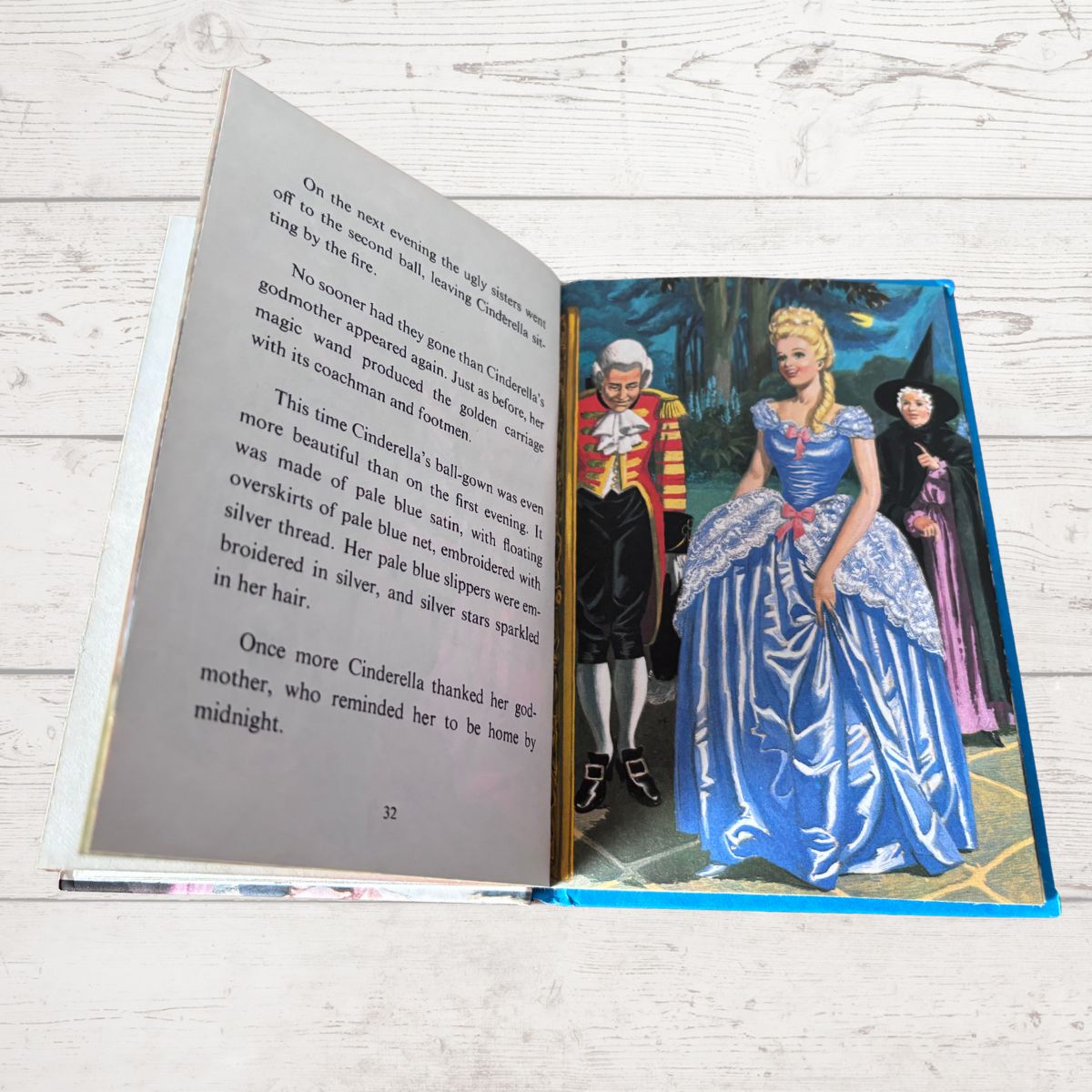 An open children's book lying on a white wood surface, showing a colorful illustration of Cinderella on the right page and text on the left page.