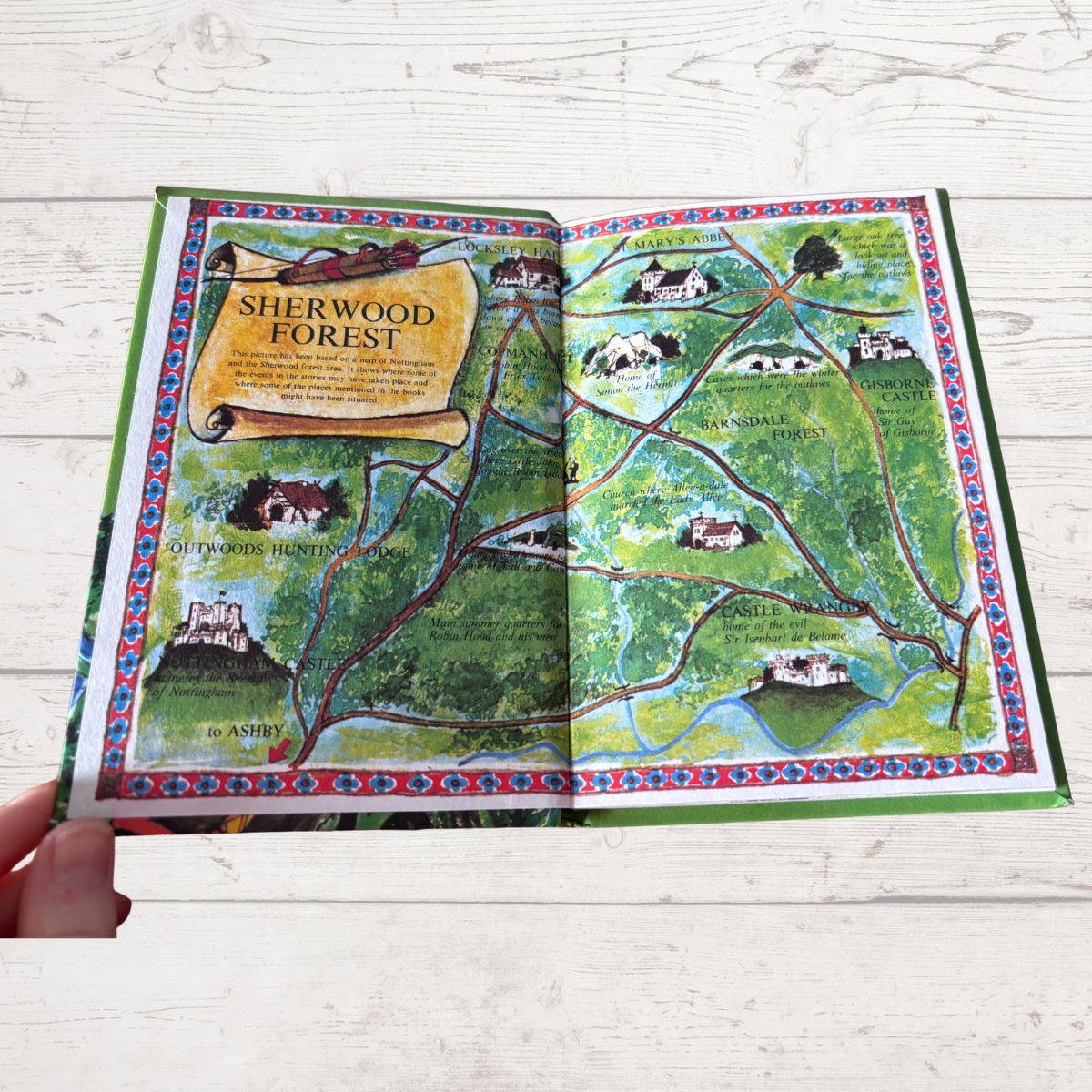 An open illustrated book titled 'Sherwood Forest' showing a colorful map with trees, houses, and winding paths, photographed on a light wood surface with a hand holding the left edge.
