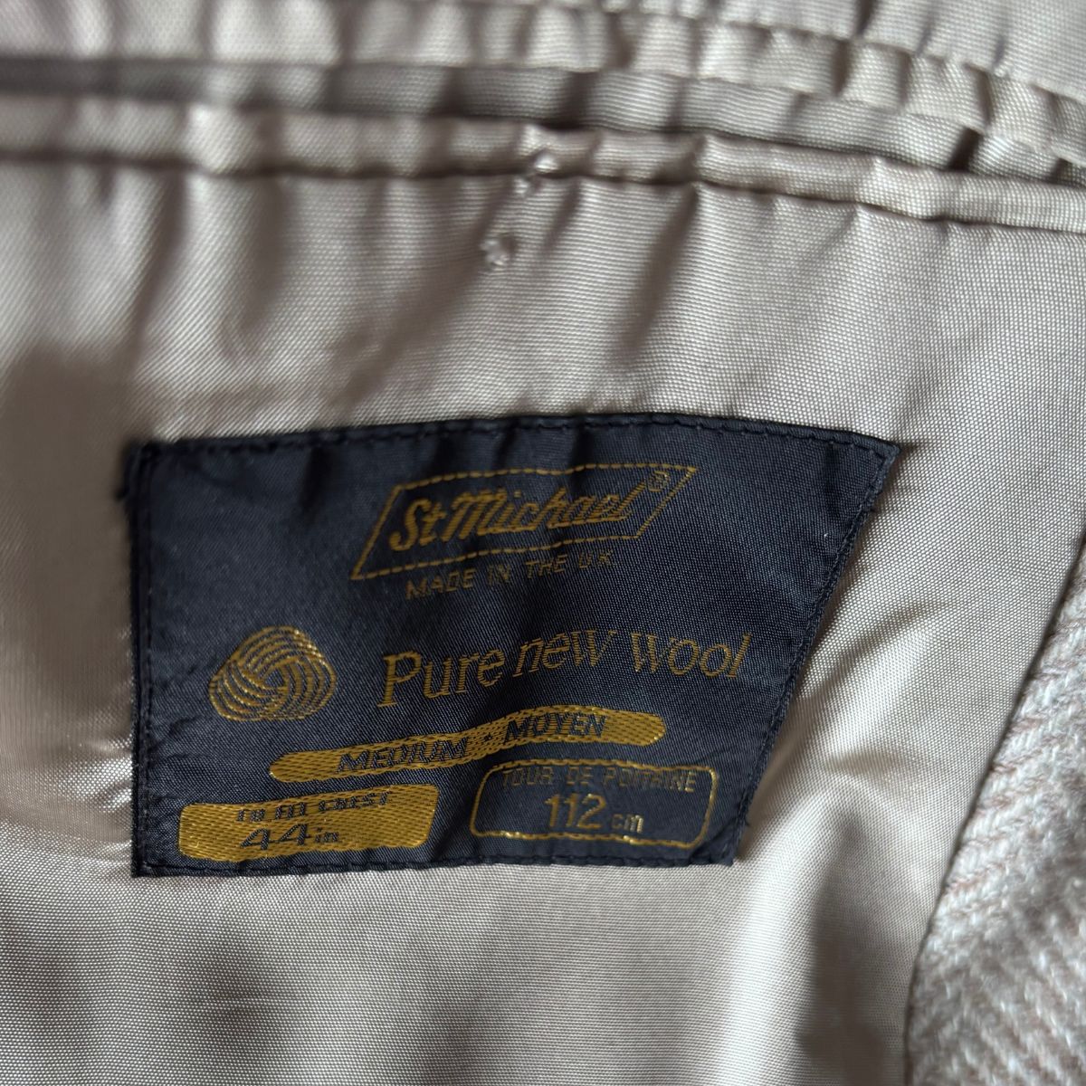 Close-up of a garment's interior label reading 'St. Michael's' and 'Pure new wool' in gold embroidery on a dark navy patch, with light beige fabric surrounding it.